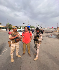 Un uomo con una camicia rossa è in piedi tra due soldati in tenuta militare, ognuno con un fucile in mano, su una strada sotto un cielo nuvoloso.