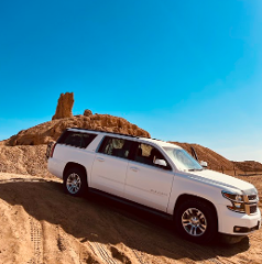 Un SUV bianco è parcheggiato su una collina sabbiosa con formazioni rocciose sotto un cielo azzurro e limpido.