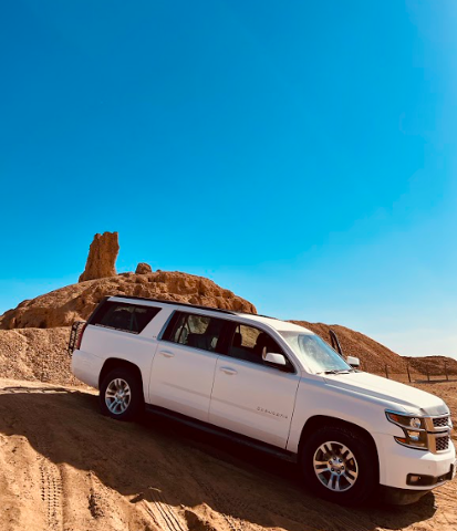A white Chevrolet Suburban SUV is parked on a sandy hill under a clear blue sky with rocky desert formations in the background.