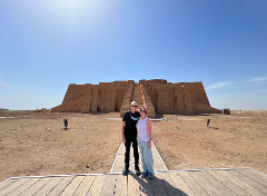 A couple poses in front of the ancient Ziggurat of Ur, a large stepped pyramid made of mud brick, under a clear blue sky.