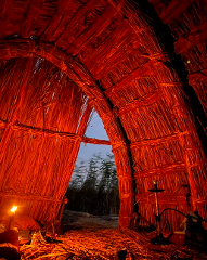 A straw hut interior is illuminated by a candle and fire, with a hookah and a view of tall grass through the opening.