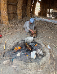 An elderly man wearing a headscarf cooks chicken over an open fire inside a straw hut.