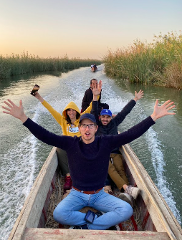 A group of five people are joyfully riding in a wooden boat through a waterway surrounded by tall reeds, with another boat visible in the distance under a bright sky.