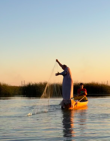 Two people are in a small boat on a river, with one man throwing a fishing net into the water as the sun sets.