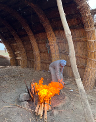 A person in traditional clothing tends to a blazing fire in front of a hut made of reeds.