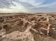 The image captures an archaeological site with the remnants of ancient mud-brick walls under a cloudy sky.