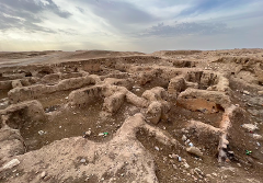 Dieses Bild zeigt eine Luftaufnahme einer archäologischen Stätte mit einem Netzwerk aus bröckelnden, erodierten Mauern und Strukturen aus Erde oder Lehm inmitten einer kargen Landschaft.