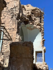 Das Bild zeigt ein teilweise zerstörtes Gebäude mit bröckelnden Mauern, einem gewölbten Eingang und einem Fenster mit kunstvollem Schmiedeeisen vor einem klaren blauen Himmel.