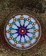 An ornate stained glass dome with a central blue star surrounded by turquoise and gold geometric patterns, set within a reflective, patterned ceiling.