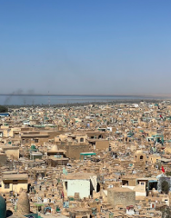 An aerial view shows a densely packed, sun-baked urban landscape leading to a hazy body of water under a clear, blue sky.