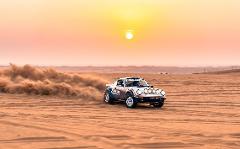 A Porsche rally car kicks up sand as it races across a desert landscape under a hazy, golden sun.
