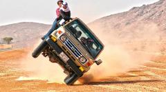 A group of people are recklessly riding a Land Cruiser, with two men sitting on the hood while the car is performing a wheelie in the desert.