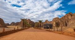 A dirt road leads through a desert landscape with rocky mountains, palm trees, and a small building under a partly cloudy sky.