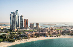 Aerial view of Abu Dhabi skyline featuring Etihad Towers, Emirates Palace, and other buildings along the coast on a sunny day.