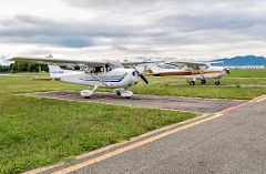 Two small planes are parked on the tarmac of a grassy airfield under a cloudy sky.