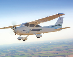 A white Cessna Skyhawk airplane soars through the sky, its propeller blurred in motion, with a landscape of fields visible below.