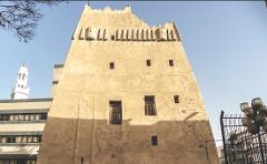 A tall, beige, fortress-like building stands prominently against a blue sky, with a modern building and a minaret visible in the background.