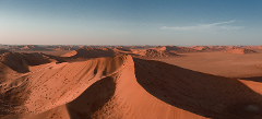 An aerial shot captures a vast desert landscape with rolling sand dunes under a clear blue sky, casting shadows that highlight the texture of the dunes.