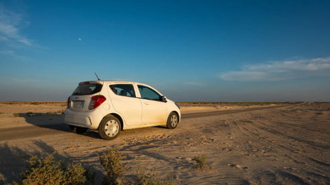 A white car is parked on a sandy road in a desert landscape under a clear blue sky with a faint moon.