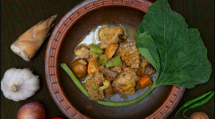 A hearty stew with meat, potatoes, carrots, and greens is served in a rustic bowl with bamboo shoots, garlic, onion, and peppers alongside.