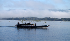 A long boat speeds across a calm body of water with two people aboard, against a backdrop of forested hills obscured by morning fog.