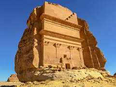 This image shows the Qasr al-Farid tomb, a large Nabataean sandstone structure carved into a rock outcrop in Mada'in Saleh, Saudi Arabia, under a clear blue sky.