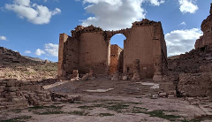 A weathered, crumbling stone temple with an arched gateway stands against a backdrop of arid hills under a partly cloudy sky.