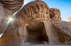 A rock-cut tomb, likely Nabataean, with a sunburst shining through a gap in the rock formations above.