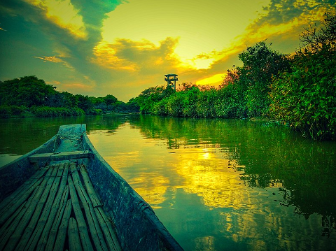 A boat floats on a river surrounded by green foliage under a sunset sky with a watchtower in the distance.