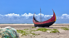 A vibrant, weathered fishing boat rests on a sandy beach under a cloudy blue sky, with scattered green plants and a fishing net in the foreground.