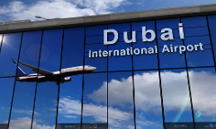 A passenger plane flies in front of the Dubai International Airport windows, reflecting the sky and clouds.
