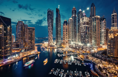This is a vibrant night scene of Dubai Marina, showcasing towering skyscrapers illuminated against the twilight sky, with boats docked in the harbor reflecting the city lights.