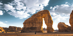 A solitary figure stands before the majestic Elephant Rock in AlUla, Saudi Arabia, under a sky filled with billowing clouds.