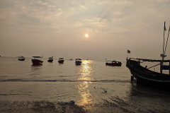 A serene sunset bathes the sea in golden light, illuminating several boats gently floating on the water and a larger boat resting on the shore.
