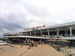 The image shows a busy exterior view of Hazrat Shahjalal International Airport Terminal-2 in Dhaka, Bangladesh, with people gathered outside the building under an overcast sky.