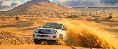 A white SUV car is driving through a vast desert, kicking up sand as it navigates the dunes, with a mountain in the background.