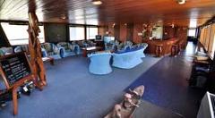 Hier ist eine Beschreibung des Bildes: Der Innenbereich des Loungebereichs des Kreuzfahrtschiffs Sepik Spirit verfügt über bequeme Sitzgelegenheiten, eine Bar und Holzdekor, was eine gemütliche und einladende Atmosphäre schafft.