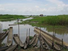 A serene river scene with several dugout canoes resting on the shore, lush greenery on both sides, and a small thatched-roof hut on stilts in the distance.