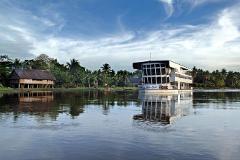 Eine ruhige Szene des Sepik-Flusses mit einem modernen Kreuzfahrtschiff, der Sepik Spirit, im Kontrast zu einem traditionellen Pfahlhaus am üppigen Flussufer unter einem teilweise bewölkten Himmel.
