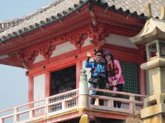 A smiling man waves while standing on a balcony with a woman in front of a red and white Japanese building with a tiled roof.