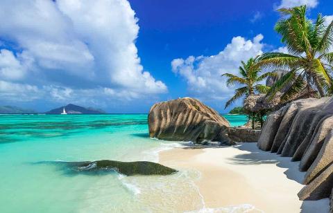 This idyllic tropical beach scene features turquoise waters, white sand, granite boulders, palm trees, and a clear blue sky with fluffy clouds.
