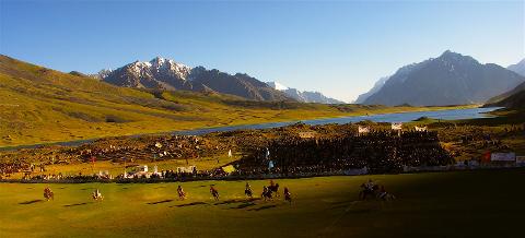 A polo match is in progress on a green field, with mountains and a lake in the background and a large crowd of spectators watching.