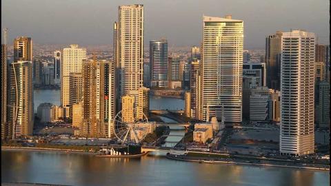 A cityscape with tall buildings, a body of water in the foreground, and a Ferris wheel near the center.