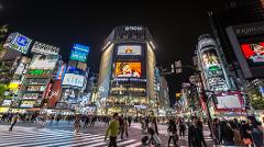 This is a vibrant night scene in Shibuya, Tokyo, filled with bustling crowds crossing the street under a dazzling array of illuminated billboards and building-sized video screens.