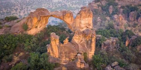 Full Day Tour To Siby And Bamako A natural sandstone arch bridges a gap between rocky outcrops covered in lush green vegetation.