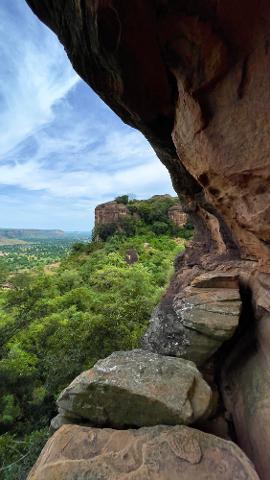 A scenic view of a lush green landscape and towering cliffs under a partly cloudy sky, framed by the dark, rocky overhang of a cave.