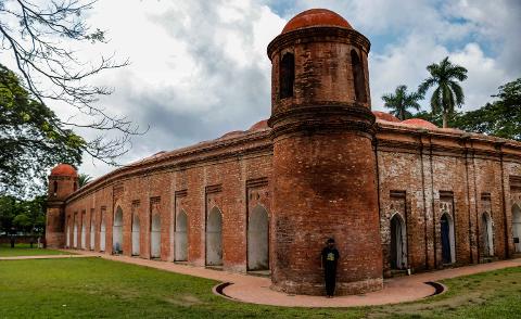 A long, red brick building with arches and a tower stands in a grassy field under a cloudy sky, with a person standing against the tower.