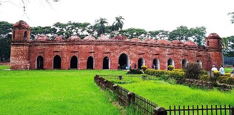 A brick building with multiple arches and domes sits on a lush green lawn, framed by trees and a low fence.
