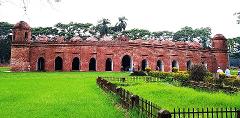 A brick building with multiple arches and domes sits on a lush green lawn, framed by trees and a low fence.