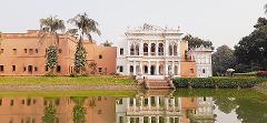 A serene reflection of the Sonargaon Panam City architecture on the water, showcasing a blend of brick and ornate white buildings surrounded by lush greenery.